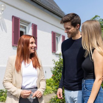 Immobilienmaklerin zeigt jungem Paar ein Haus von außen