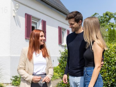 Immobilienmaklerin zeigt jungem Paar ein Haus von außen